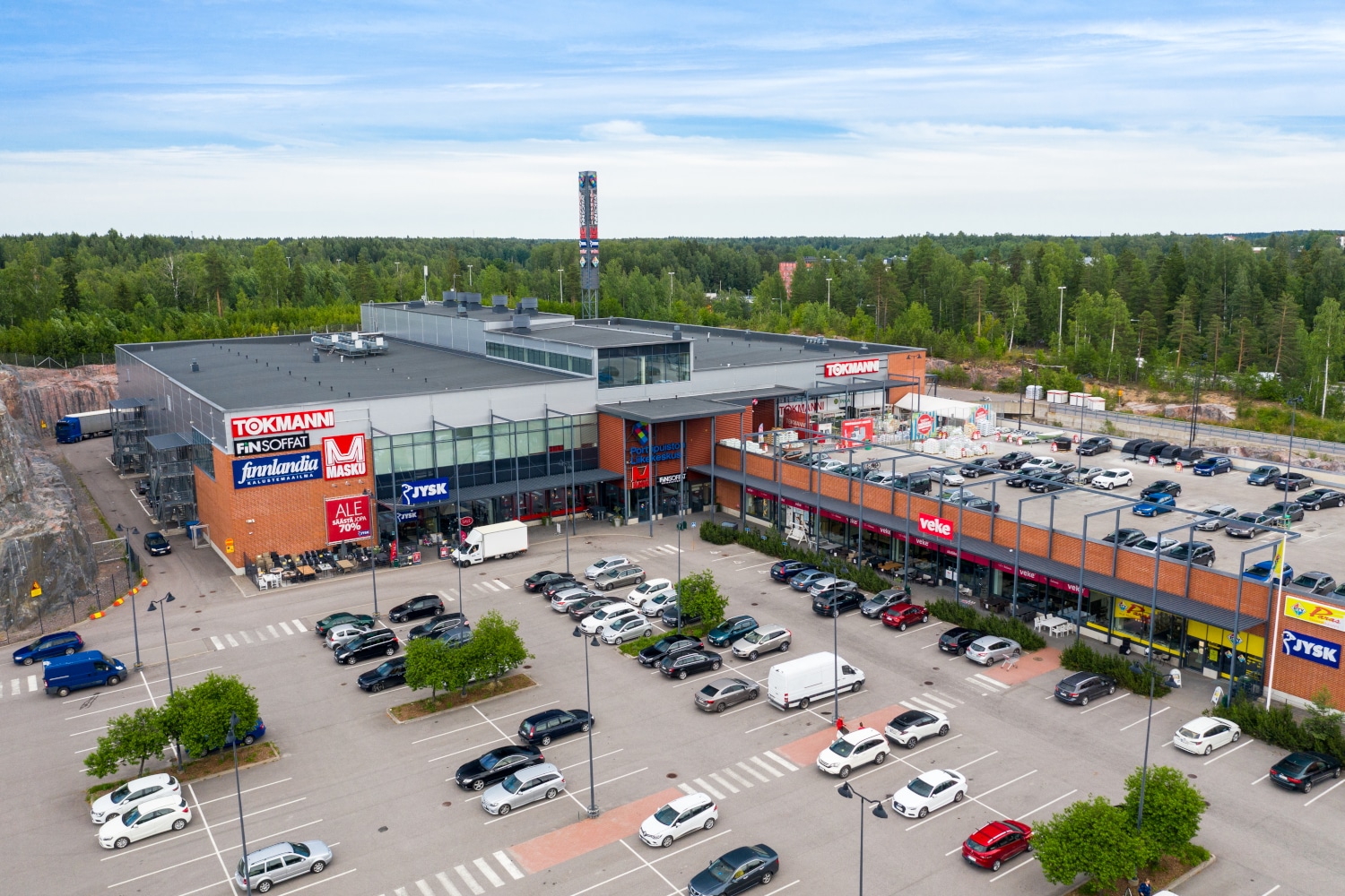 Ilmakuva kauppakeskuksesta osoitteessa Porttisuontie 4, L-muotoinen talo jonka edessä autoja pysäköintipaikalla.